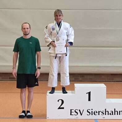 Drei Personen stehen auf einem Siegertreppchen in einer Sporthalle, einer h&auml;lt einen Pokal, zwei tragen gr&uuml;ne T-Shirts.