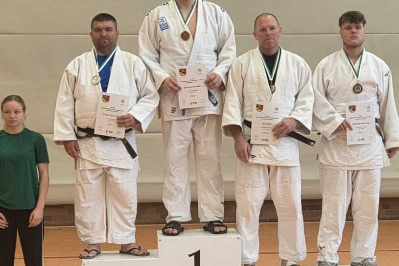 Drei Judokas in wei&szlig;en Anz&uuml;gen mit Medaillen stehen auf dem Siegertreppchen, w&auml;hrend zwei Zuschauer daneben stehen.