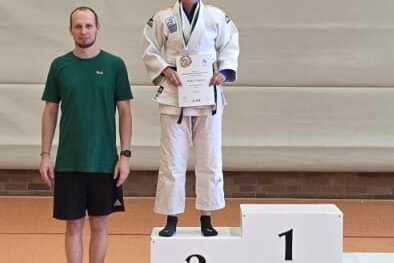 Drei Personen stehen auf einem Siegertreppchen in einer Sporthalle, einer h&auml;lt einen Pokal, zwei tragen gr&uuml;ne T-Shirts.