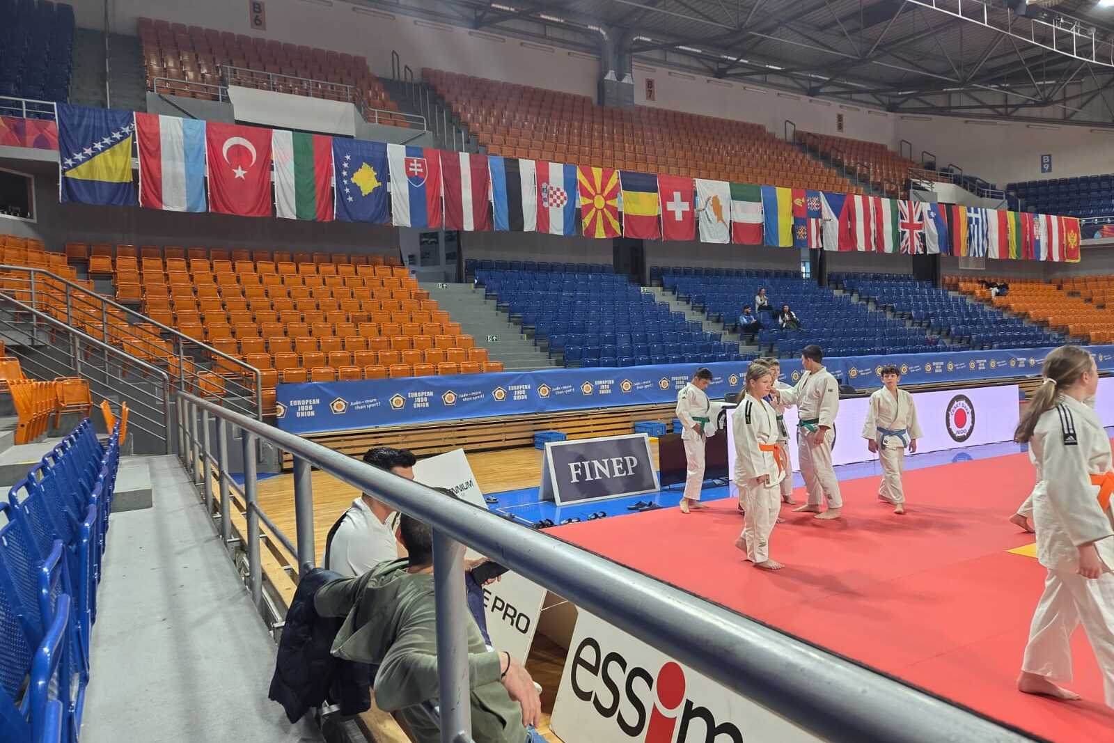 Innenansicht einer Sporthalle mit Flaggen, leeren Sitzplätzen und Teilnehmern in weißen Trainingsanzügen auf der Matte.