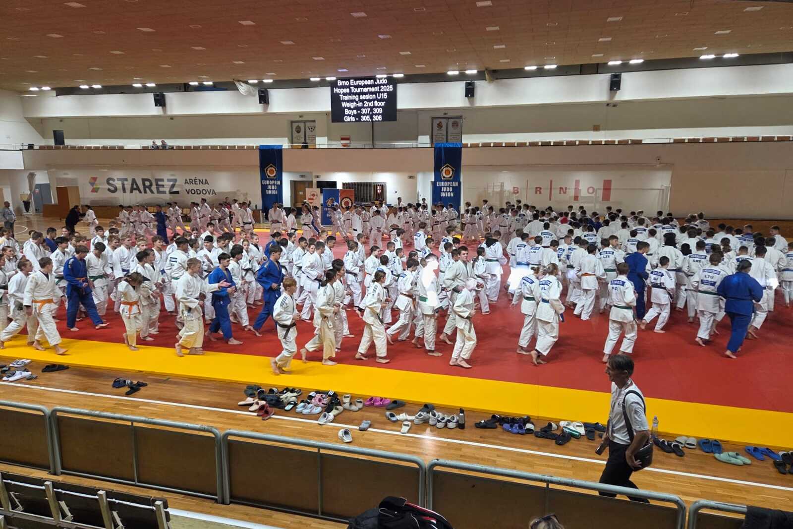 Große Gruppe von Judokas in weißen und blauen Anzügen auf einer roten Matte während eines Wettkampfs in einer Sporthalle.