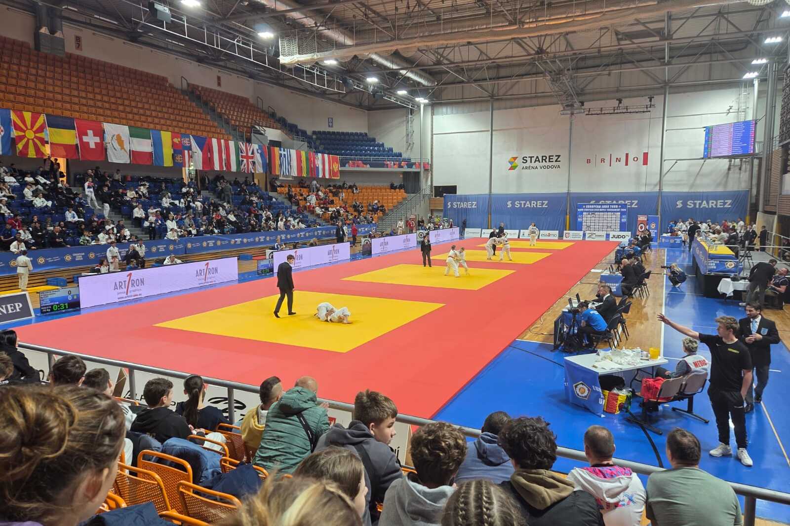 Zuschauer in einer Sporthalle beobachten Wettkämpfe auf zwei Judomatten, umgeben von Flaggen und Werbebannern.