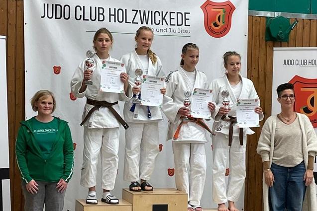 F&uuml;nf M&auml;dchen in Judogew&auml;ndern stehen auf dem Treppchen, halten Urkunden und l&auml;cheln vor einem Banner des Judo Clubs.
