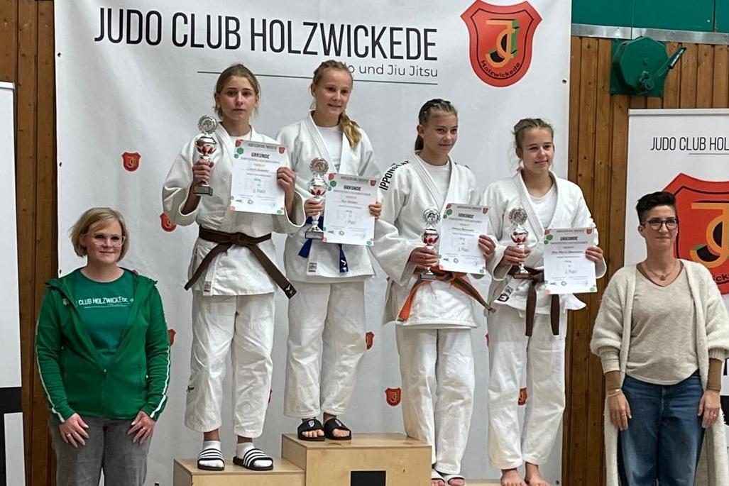 Fünf Mädchen in Judogewändern stehen auf dem Treppchen, halten Urkunden und lächeln vor einem Banner des Judo Clubs.