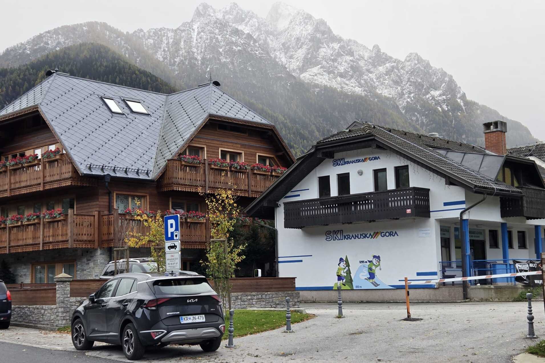 Zwei alpine Geb&auml;ude mit Holzverkleidung, neben einem Parkplatz, vor einer bergigen Kulisse. Grauer Himmel.