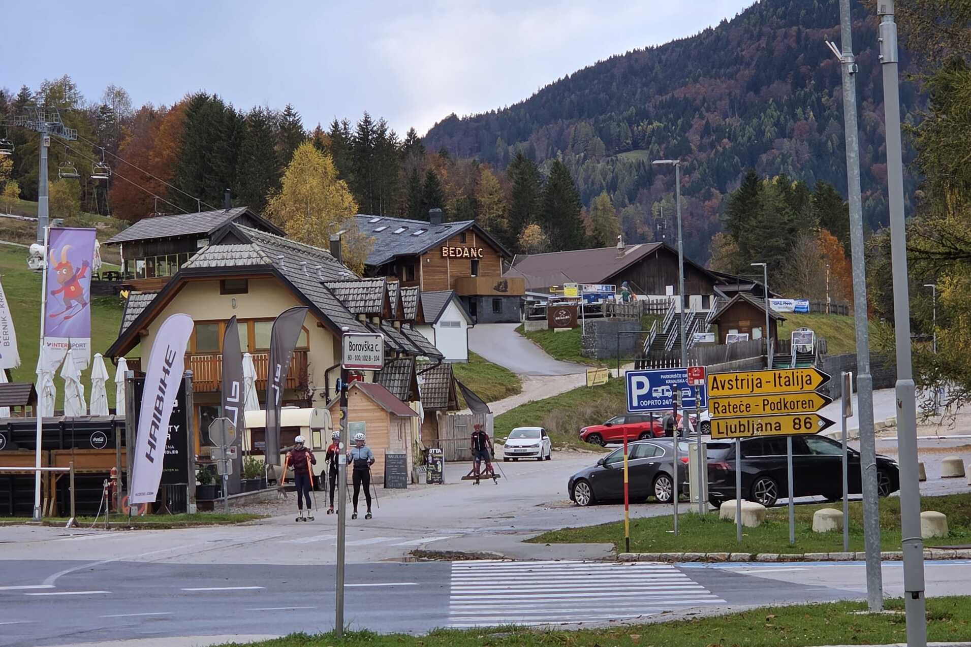 Blick auf eine Bergh&uuml;ttenlandschaft mit Skifahrern auf einem Weg, umgeben von B&auml;umen und Bergen im Hintergrund.