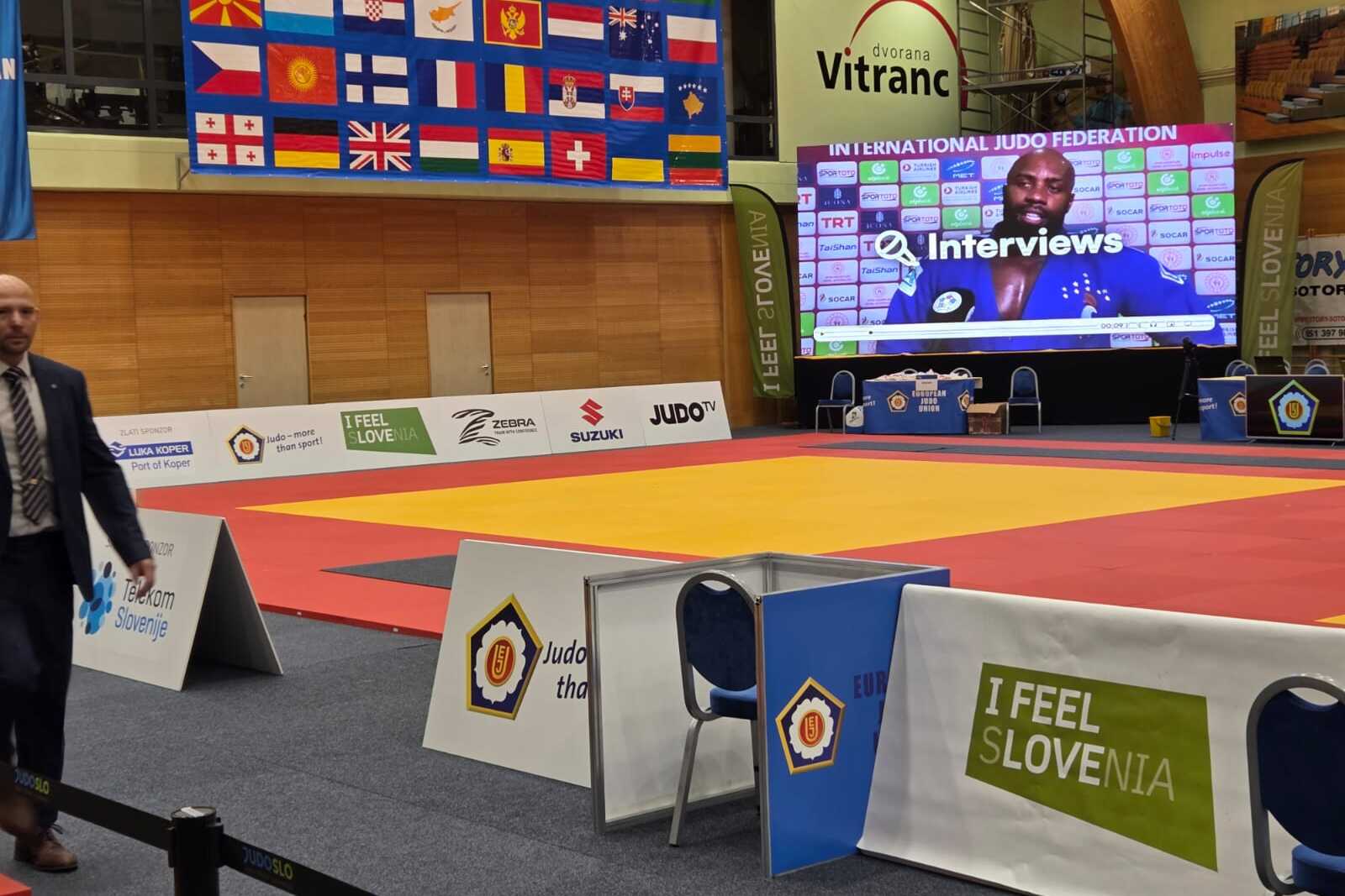 Leere Judohalle mit bunten Matten, einem gro&szlig;en Bildschirm und Flaggen im Hintergrund; ein Mann geht auf einen Stuhl zu.