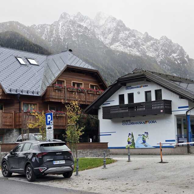 Zwei alpine Geb&auml;ude mit Holzverkleidung, neben einem Parkplatz, vor einer bergigen Kulisse. Grauer Himmel.