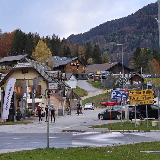 Blick auf eine Bergh&uuml;ttenlandschaft mit Skifahrern auf einem Weg, umgeben von B&auml;umen und Bergen im Hintergrund.