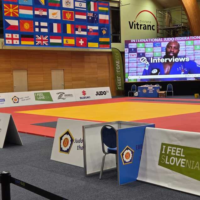Leere Judohalle mit bunten Matten, einem gro&szlig;en Bildschirm und Flaggen im Hintergrund; ein Mann geht auf einen Stuhl zu.