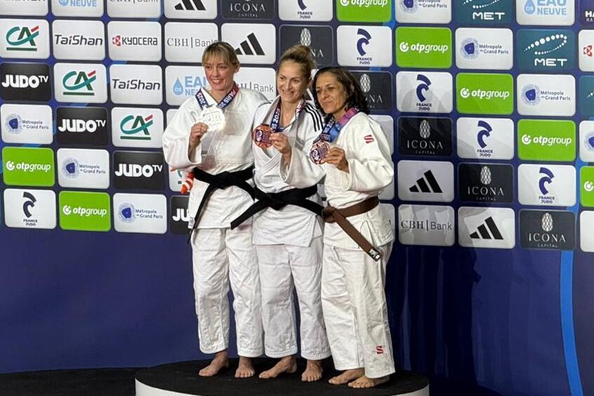 Drei Judoka in weißem Gi mit schwarzen Gürteln halten Medaillen, stehen auf einem Podest vor einer Bannerwand.