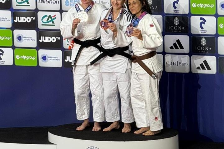 Drei Judoka in wei&szlig;em Gi mit schwarzen G&uuml;rteln halten Medaillen, stehen auf einem Podest vor einer Bannerwand.