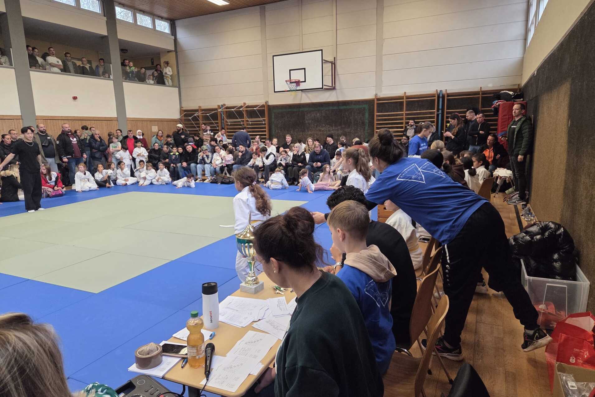 Ein Kampfsportturnier in einer Halle mit Zuschauern, Kämpfern auf einer Matte und einer Wettkampf-Umgebung.
