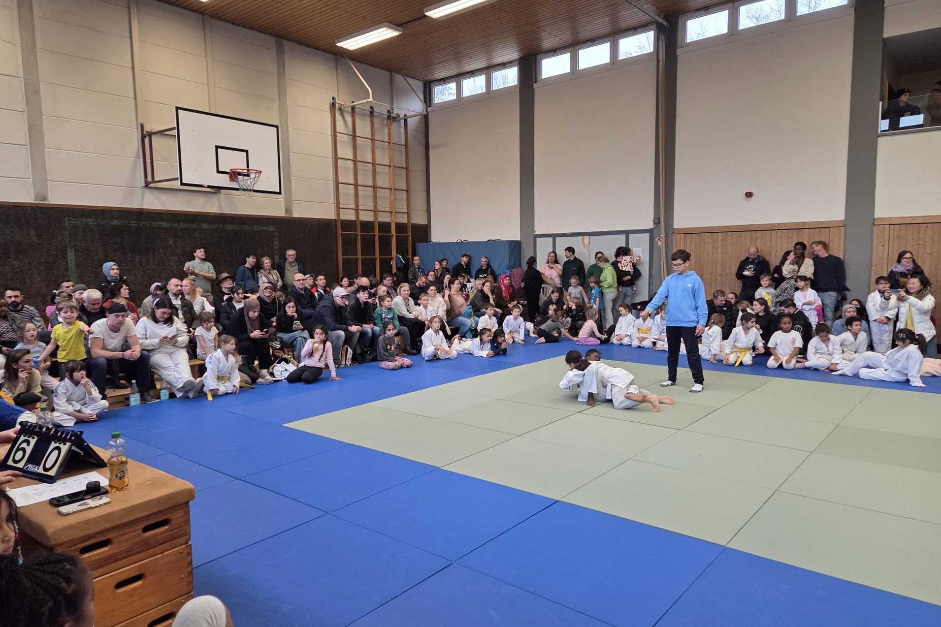 Judo-Vorführung mit Teilnehmern auf einer Matte, umgeben von Zuschauern in einer Turnhalle.