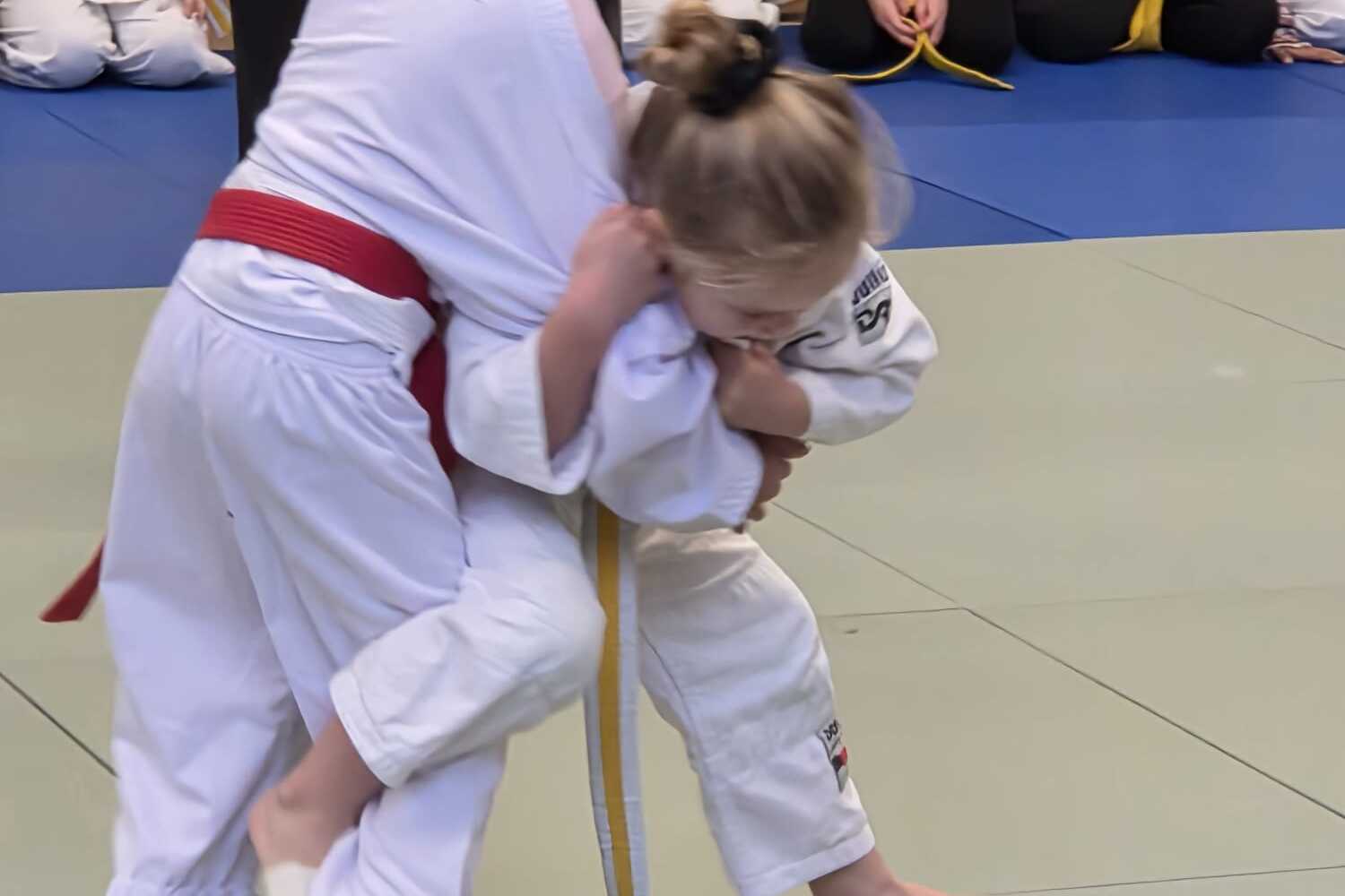 Zwei Kinder im Judokampf auf einer Matte, eines hält das andere mit einem Griff fest, Zuschauer im Hintergrund.