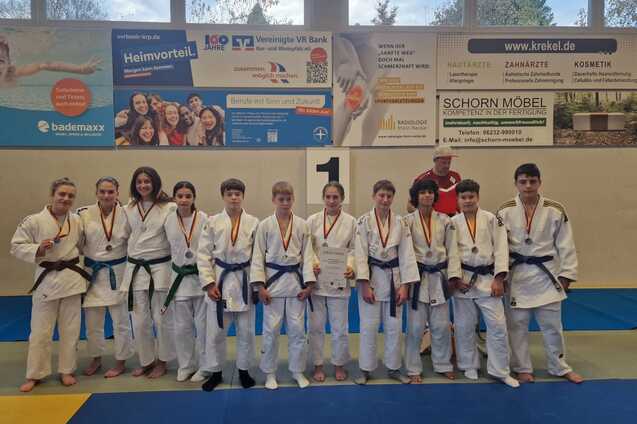 Gruppe von Judoka in Uniform mit Medaillen auf einer Judomatte, Umgebungsplakate im Hintergrund.