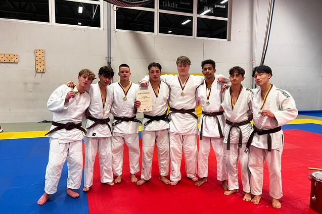 Gruppe von acht Judoka in traditionellen Anz&uuml;gen mit schwarzen G&uuml;rteln, l&auml;chelnd auf einer Judomatte.