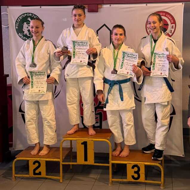 Siegerfoto von Wettk&auml;mpfern in Judoanz&uuml;gen auf einem Treppchen mit Medaillen und Urkunden, umrahmt von Betreuern.