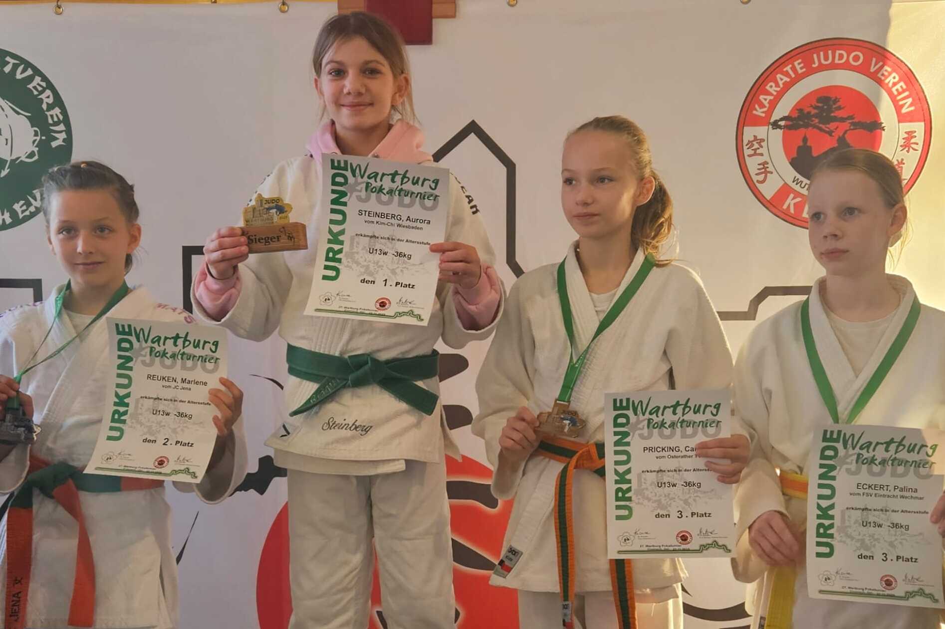 Vier junge Judoka in Gi stehen zusammen, eine auf einem Podest mit Pokal, alle halten Urkunden in der Hand.