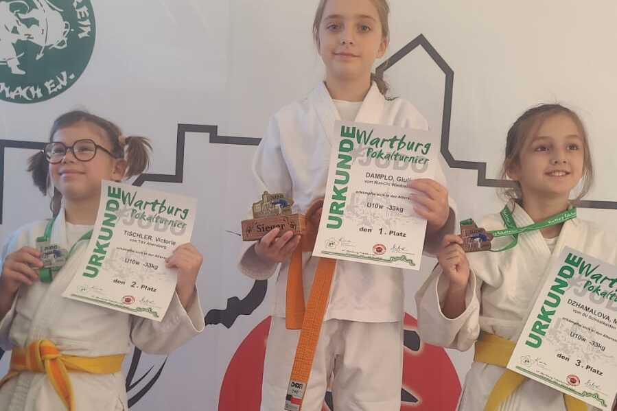 Drei Kinder in Judoanzügen stehen auf Treppchen mit Medaillen und Urkunden nach einem Wettkampf.