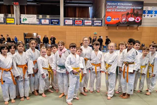 Gruppe von Kindern in wei&szlig;en Judoanz&uuml;gen mit orangen und gelben G&uuml;rteln auf einer Judo-Matte w&auml;hrend eines Wettkampfs.