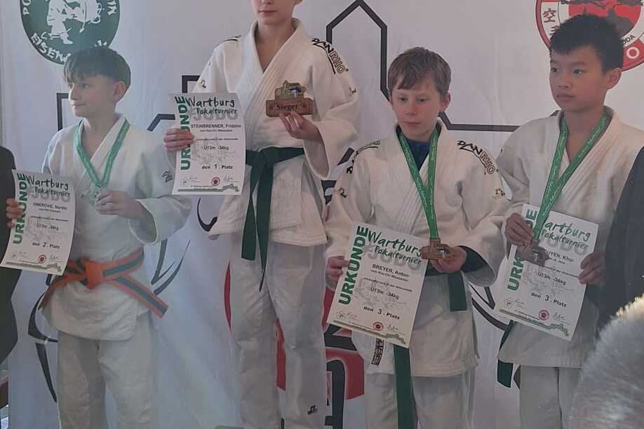 Vier Jungen in Judogis stehen auf einem Podium mit Medaillen und Urkunden, während Zuschauer im Hintergrund stehen.