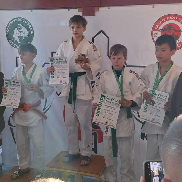 Vier Jungen in Judogis stehen auf einem Podium mit Medaillen und Urkunden, w&auml;hrend Zuschauer im Hintergrund stehen.