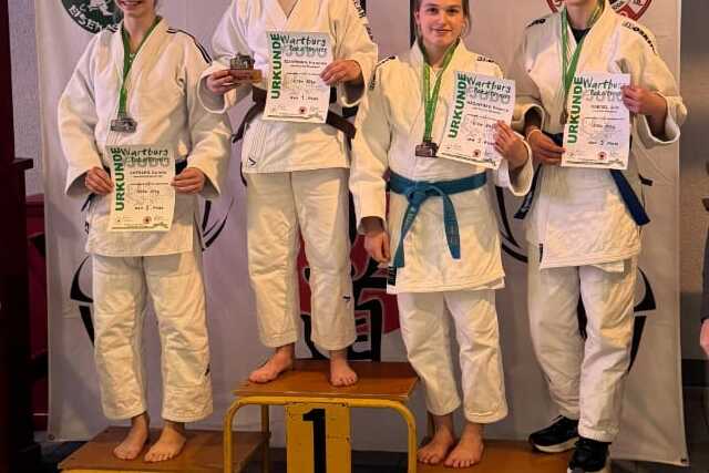 Siegerfoto von Wettkämpfern in Judoanzügen auf einem Treppchen mit Medaillen und Urkunden, umrahmt von Betreuern.