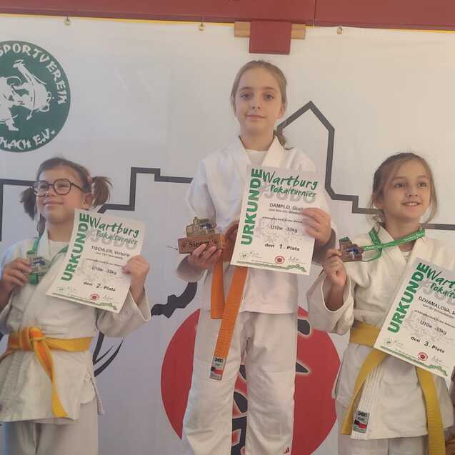 Drei Kinder in Judoanz&uuml;gen stehen auf Treppchen mit Medaillen und Urkunden nach einem Wettkampf.