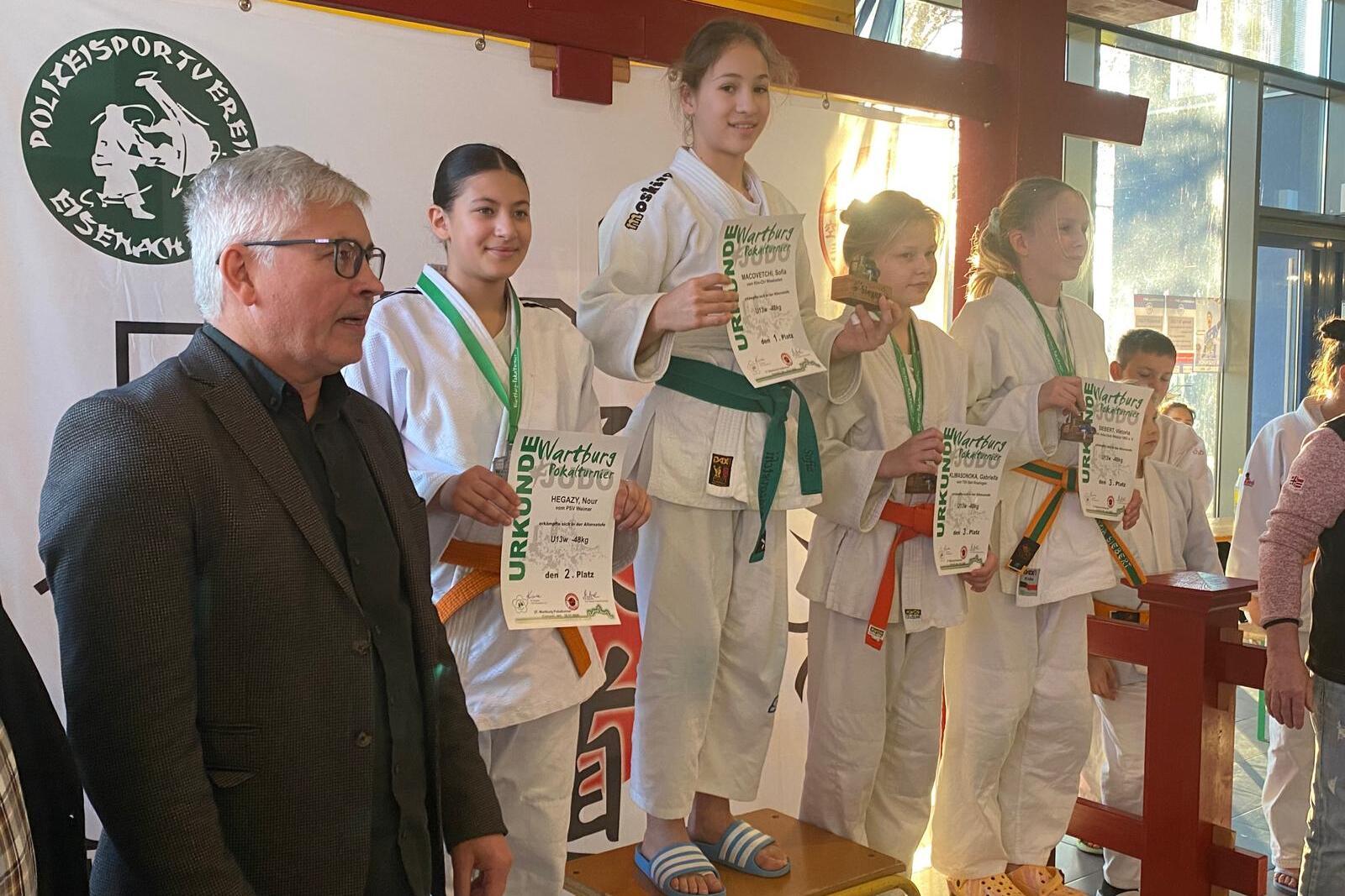 Siegerinnen stehen mit Urkunden auf dem Podium, während ein Mann in Erwachsenenkleidung zuschaut. Veranstaltungsort mit Postern.