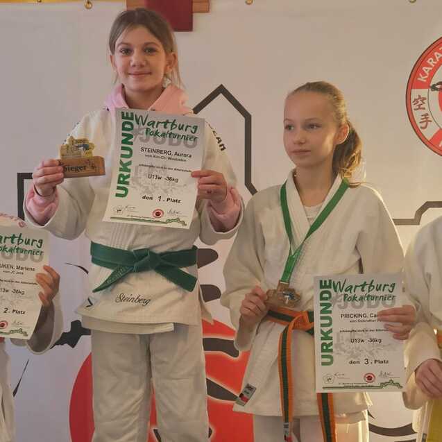 Vier junge Judoka in Gi stehen zusammen, eine auf einem Podest mit Pokal, alle halten Urkunden in der Hand.