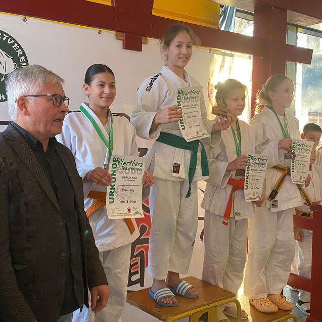 Siegerinnen stehen mit Urkunden auf dem Podium, w&auml;hrend ein Mann in Erwachsenenkleidung zuschaut. Veranstaltungsort mit Postern.