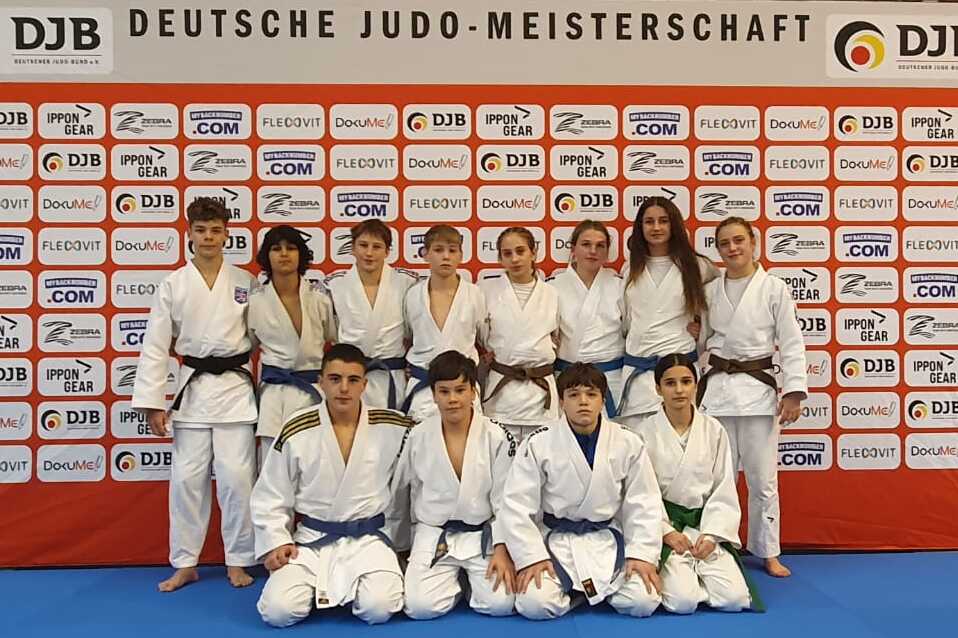 Gruppe von Judokas in weißen Anzügen vor einem Banner der Deutschen Judo-Meisterschaft, auf blauer Matte.