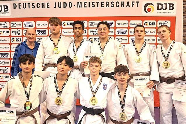 Gruppe von zehn Judokas mit Medaillen und Urkunden vor einer DJB-Wand, in Sportanz&uuml;gen auf einer blauen Matte.