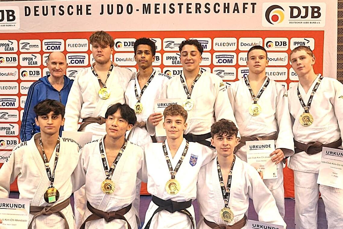 Gruppe von zehn Judokas mit Medaillen und Urkunden vor einer DJB-Wand, in Sportanzügen auf einer blauen Matte.