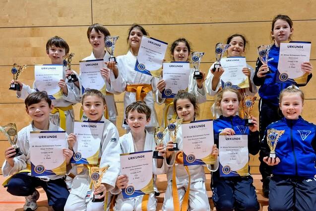 Gruppennaufnahme von zw&ouml;lf Kindern in Judo-Anz&uuml;gen mit Urkunden und Pokalen, die stolz in die Kamera l&auml;cheln.