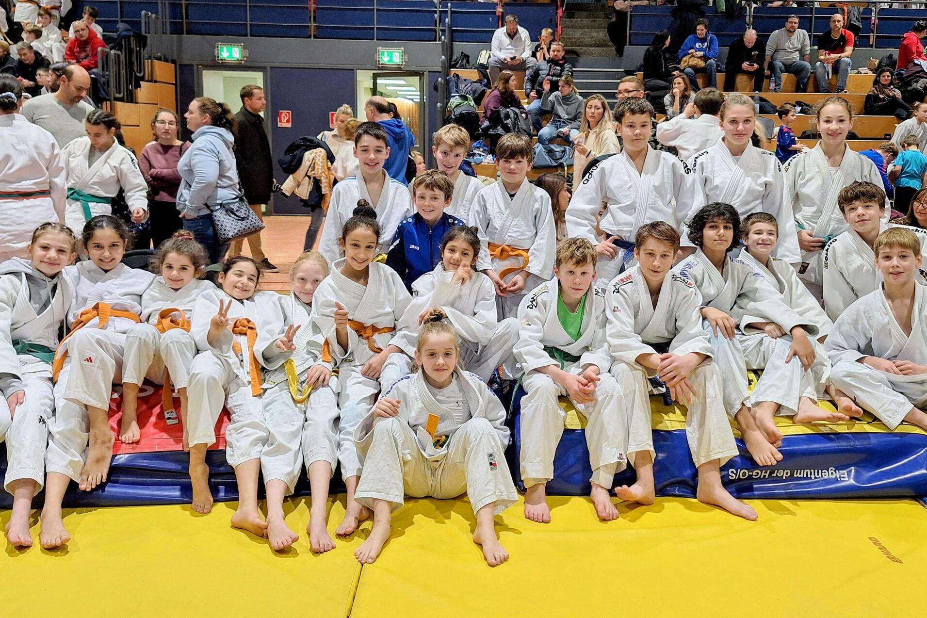 Gruppe von Jugendlichen in Judoanzügen mit verschiedenen Gürtelfarben, auf einer Matte in einer Sporthalle.