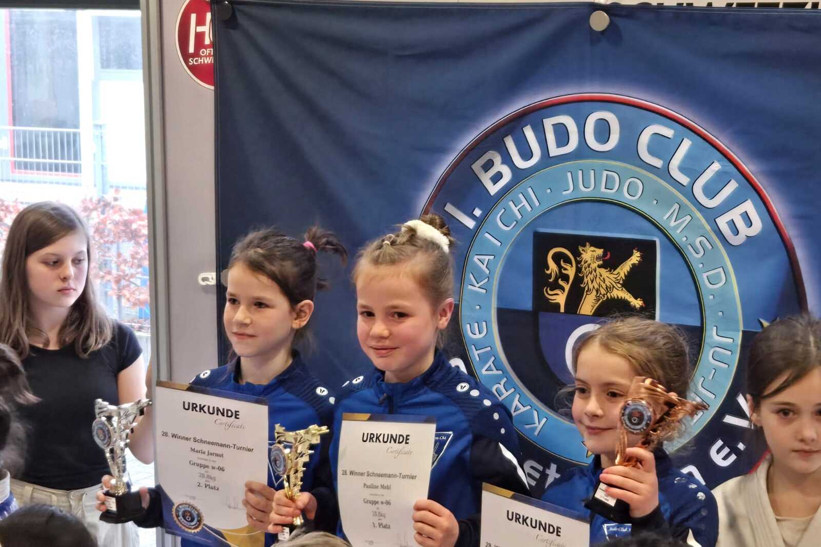 Drei Mädchen in Judoanzügen halten Urkunden und Pokale vor einem Banner des Budo Clubs.