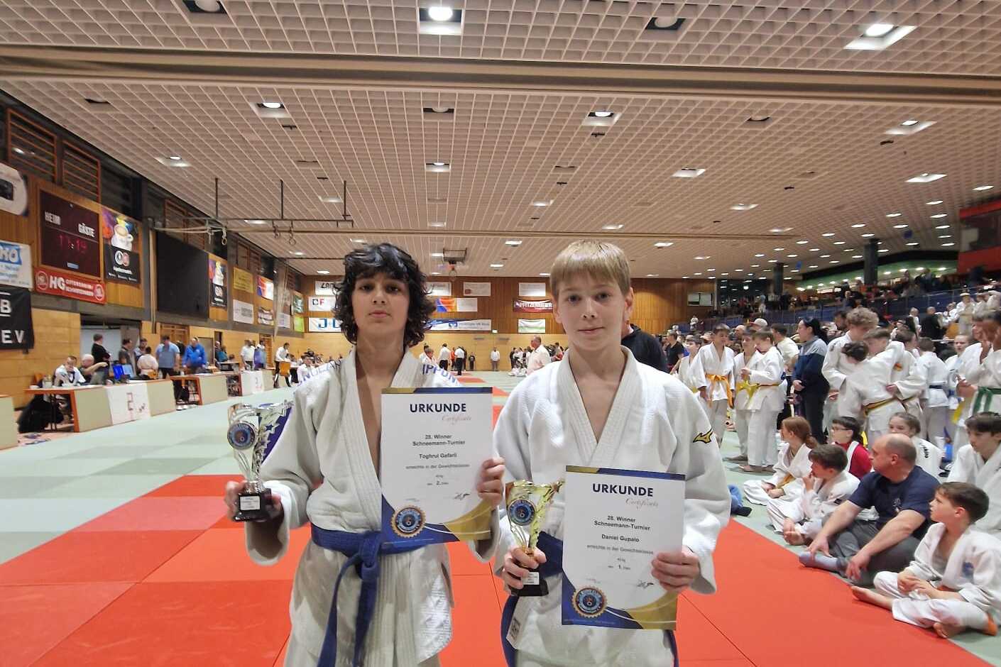 Zwei Judokas in weißen Judogis halten Urkunden und Pokale in einer Sporthalle mit Zuschauer*innen im Hintergrund.