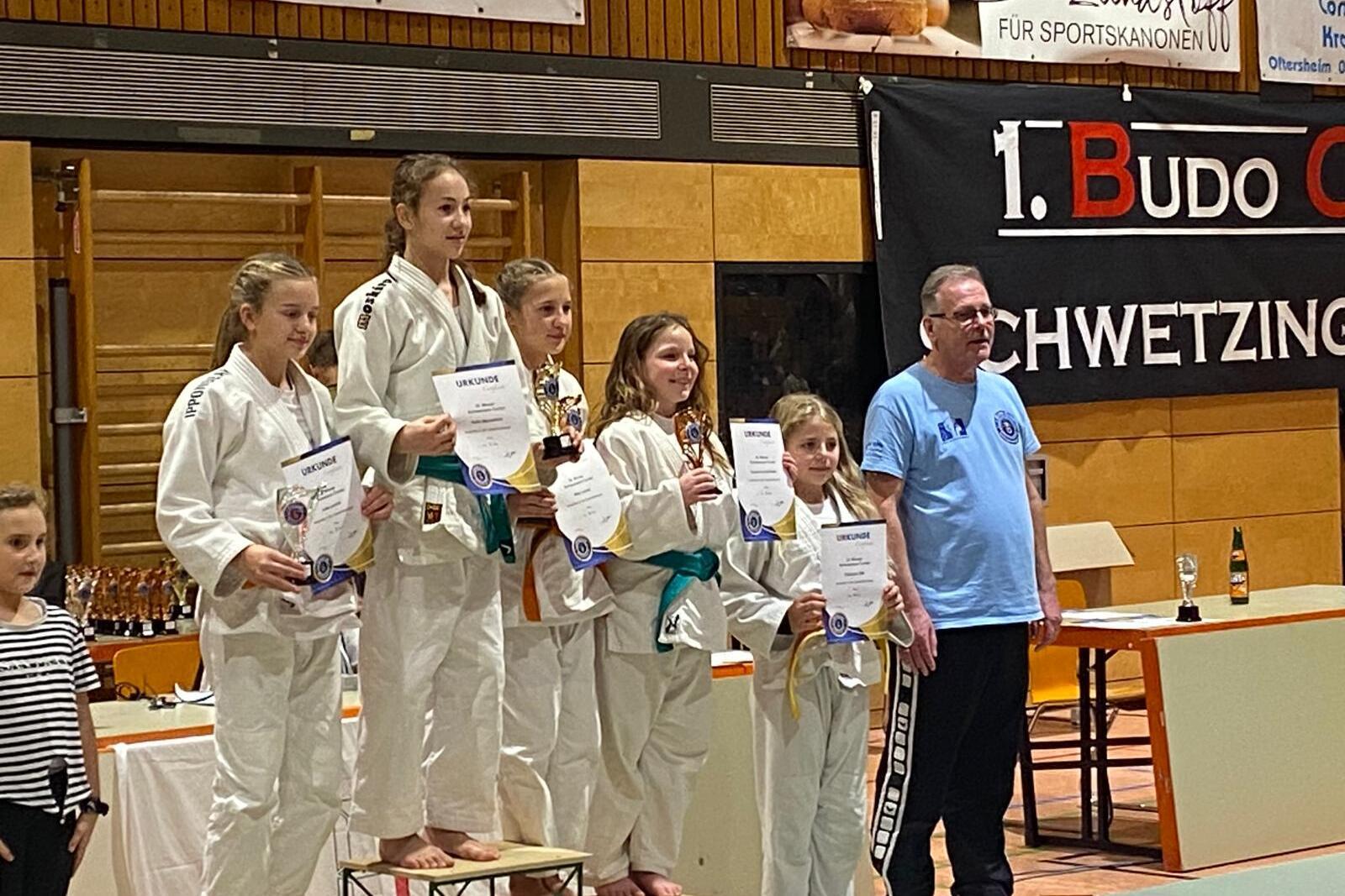 Siegerpodium mit fünf judosportlichen Kindern in Gi, die Urkunden und Pokale halten, neben einem Erwachsenen in blauem Shirt.