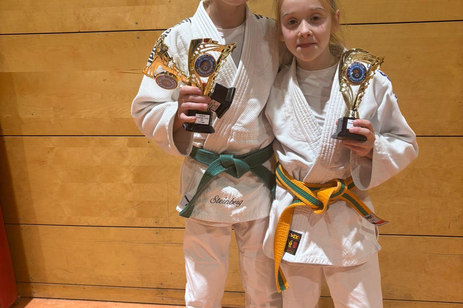Zwei Mädchen in Judogi halten Pokale nach einem Wettkampf vor einer Holzwand.