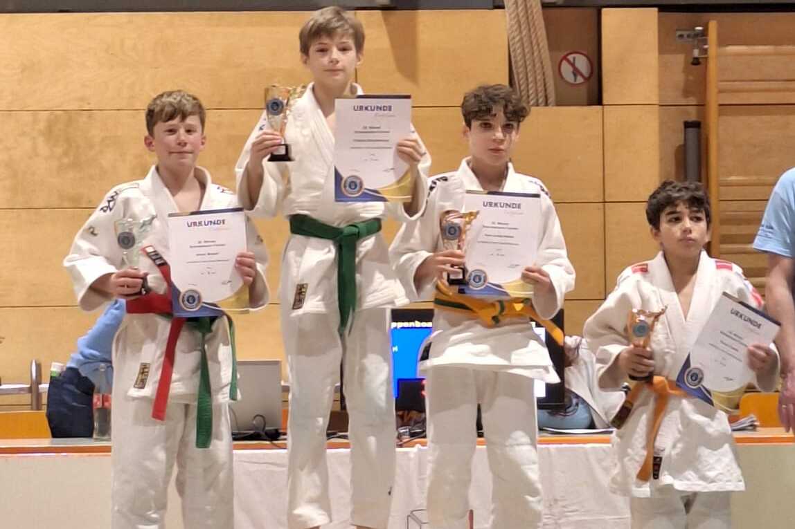 Vier Kinder in Judogi stehen auf einem Siegertreppchen und halten Pokale und Urkunden. Der Erste trägt einen grünen Gürtel.