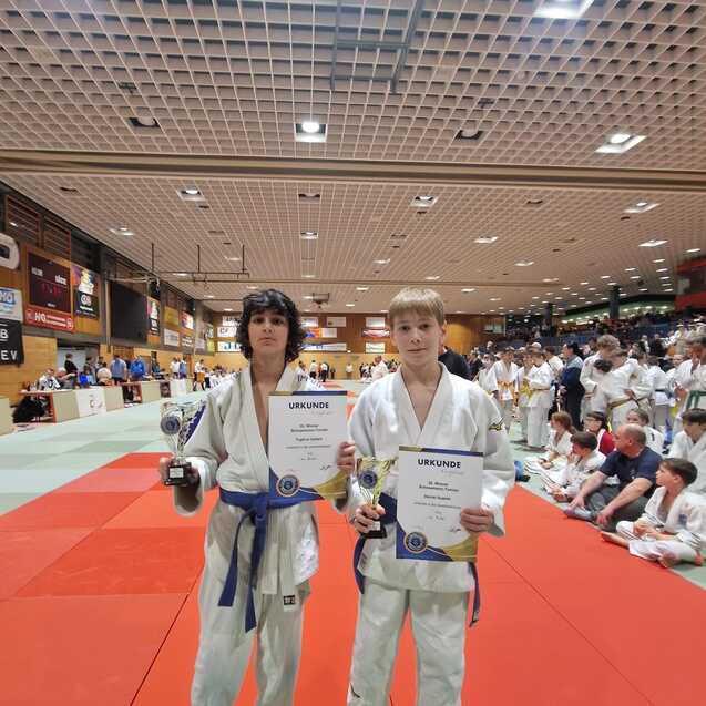 Zwei Judokas in wei&szlig;en Judogis halten Urkunden und Pokale in einer Sporthalle mit Zuschauer*innen im Hintergrund.