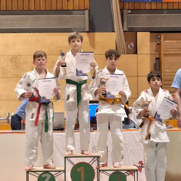 Vier Kinder in Judogi stehen auf einem Siegertreppchen und halten Pokale und Urkunden. Der Erste tr&auml;gt einen gr&uuml;nen G&uuml;rtel.