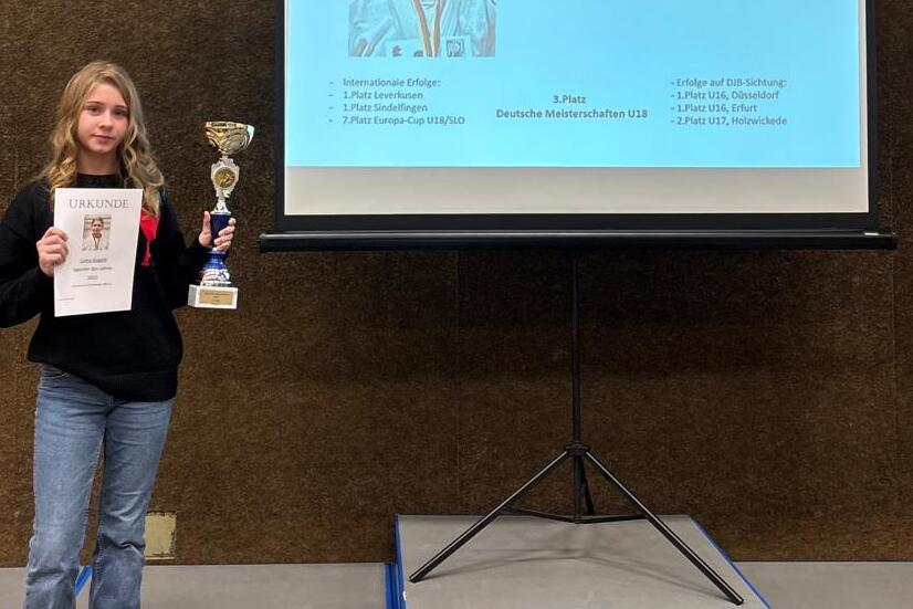 M&auml;dchen h&auml;lt eine Urkunde und einen Pokal vor einer Bildschirmpr&auml;sentation mit ihrem Namen und Foto.