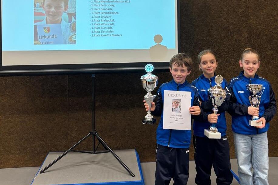 Drei Kinder stehen vor einem Pr&auml;sentationsscreen und halten Pokale sowie eine Urkunde in der Hand.