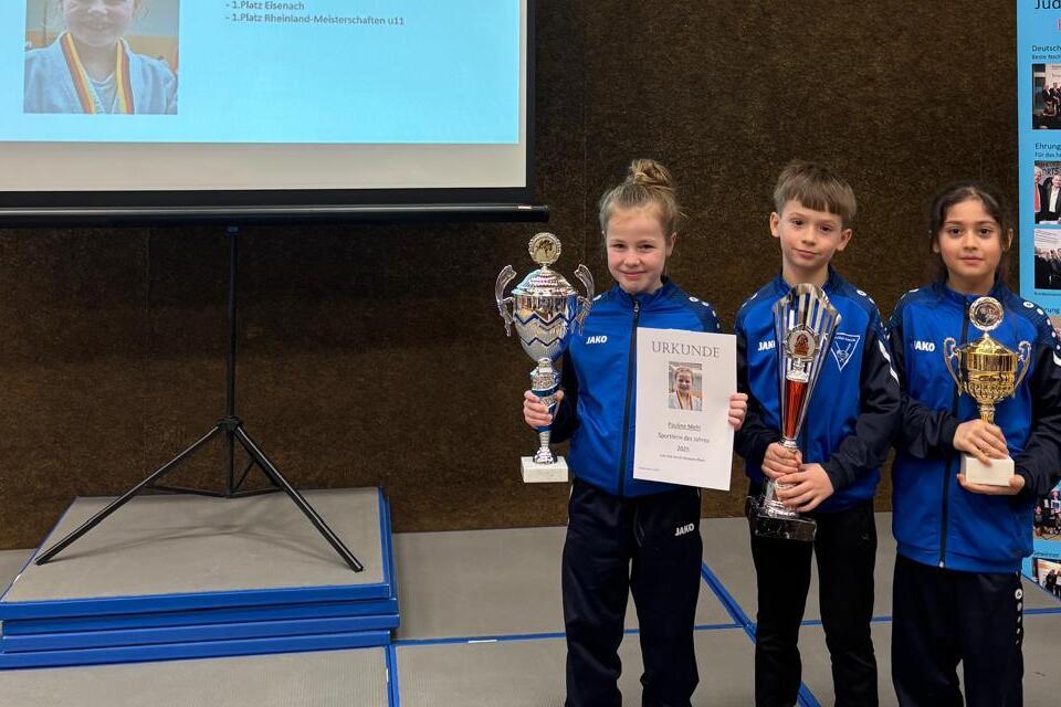 Drei Kinder stehen auf einer B&uuml;hne, halten Pokale und eine Urkunde, hinter ihnen ein Bildschirm mit Informationen.