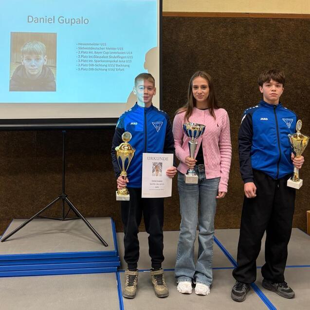 Drei Personen stehen vor einer Pr&auml;sentation, halten Pokale und Urkunden in einem Sportumfeld.