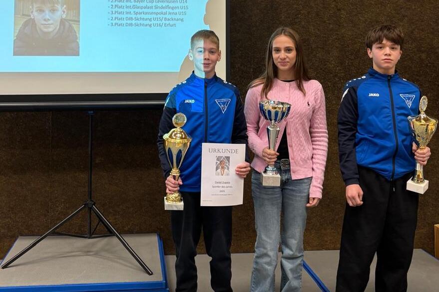 Drei Personen stehen vor einer Pr&auml;sentation, halten Pokale und Urkunden in einem Sportumfeld.