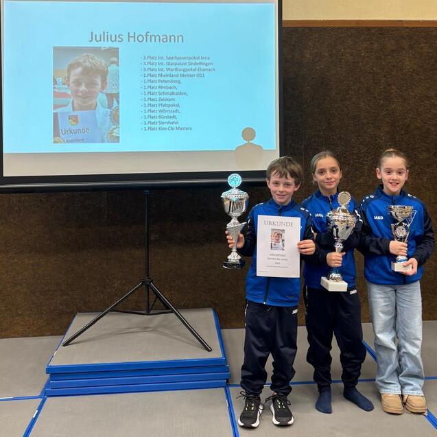 Drei Kinder stehen vor einem Pr&auml;sentationsscreen und halten Pokale sowie eine Urkunde in der Hand.